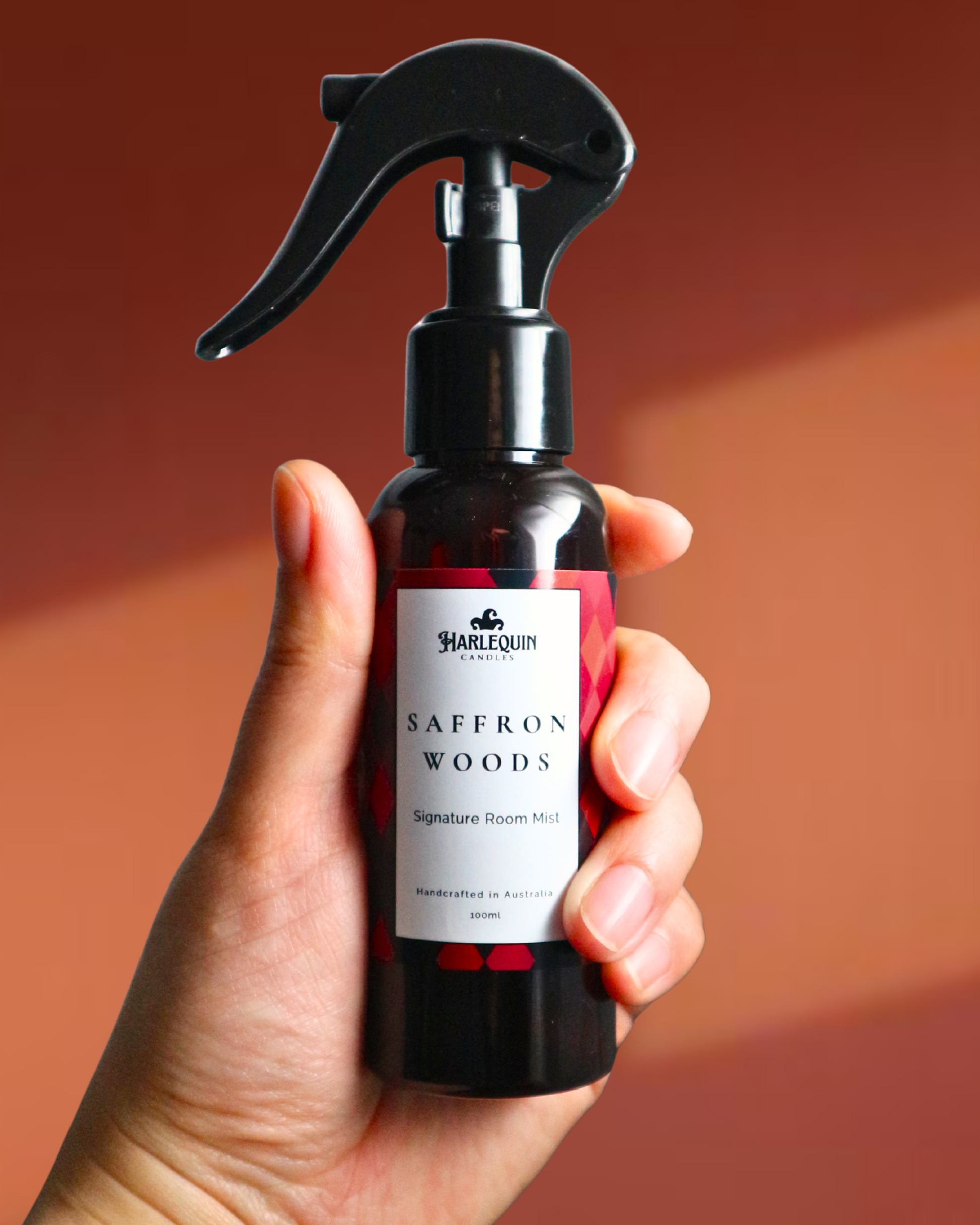 A Harlequin Candles Saffron and Sandalwood Signature Room Mist in an amber spray bottle with a black trigger cap, placed against a two-toned background orange and brown. The label clearly shows 'Harlequin Candles', 'Saffron Woods', 'Signature Room Mist', and 'Handcrafted in Australia', highlighting this enchanting Australian-made home spray.