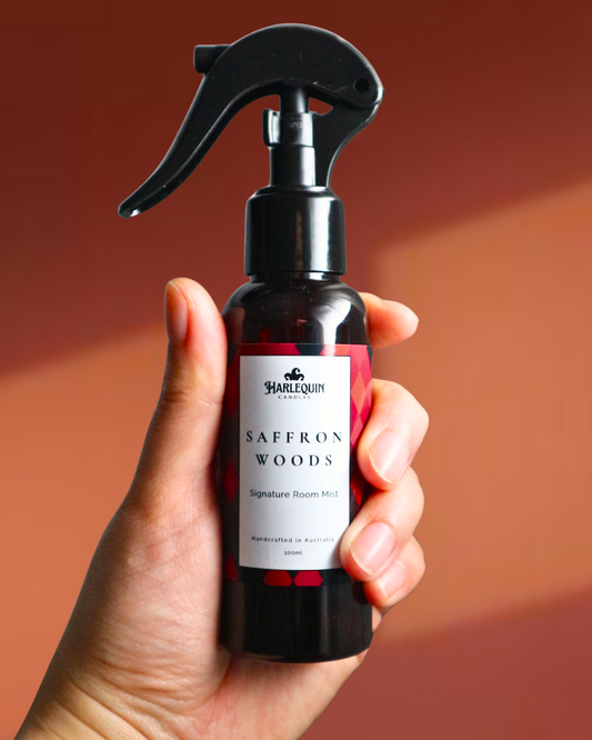 A Harlequin Candles Saffron and Sandalwood Signature Room Mist in an amber spray bottle with a black trigger cap, placed against a two-toned background orange and brown. The label clearly shows 'Harlequin Candles', 'Saffron Woods', 'Signature Room Mist', and 'Handcrafted in Australia', highlighting this enchanting Australian-made home spray.