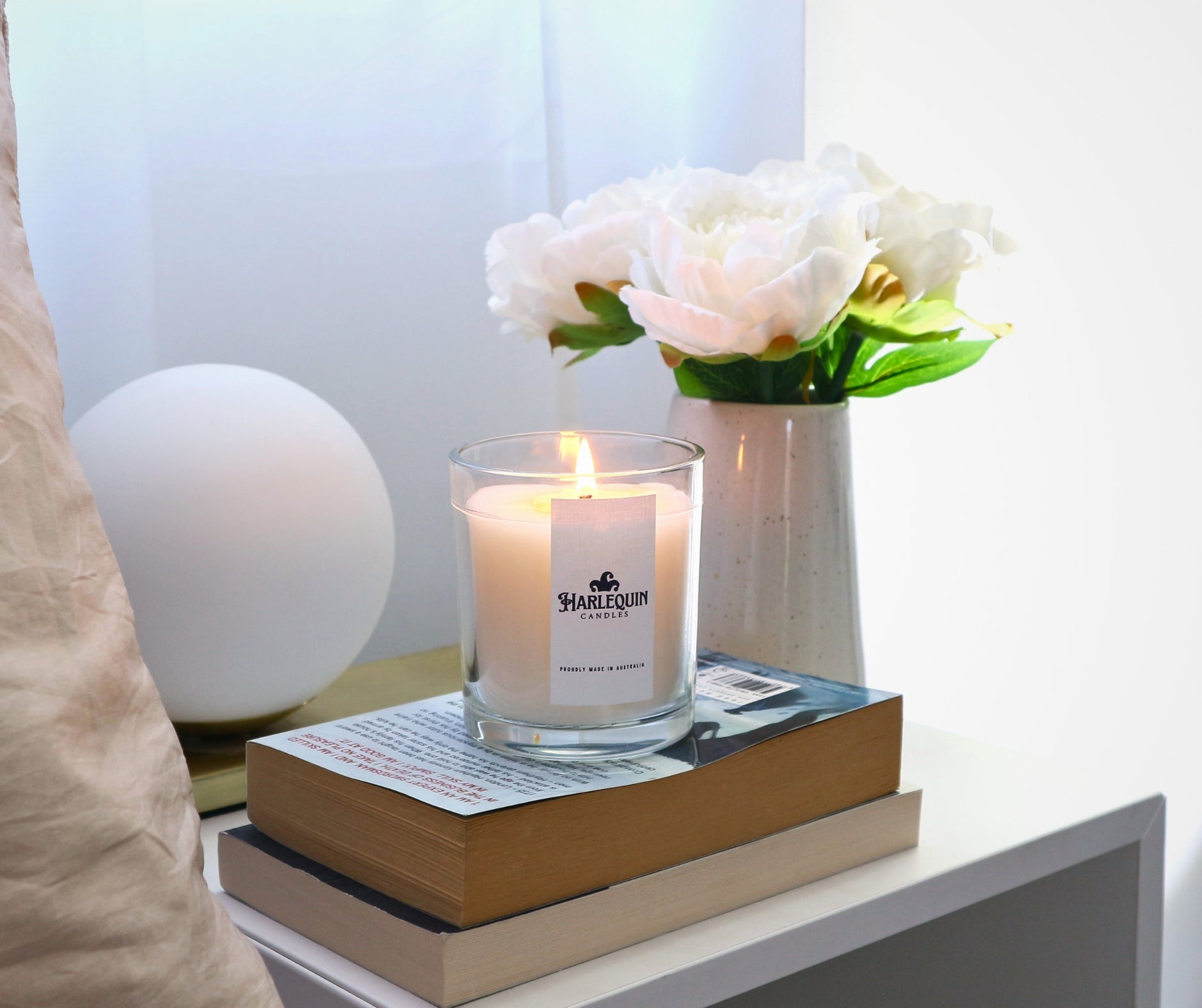 A lit Harlequin Candles soy candle in a clear glass jar with a white label reading 'PROUDLY MADE IN AUSTRALIA', resting on a stack of books on a bedside table in a bright bedroom. A spherical white lamp and a vase with white flowers complete the serene and inviting atmosphere.