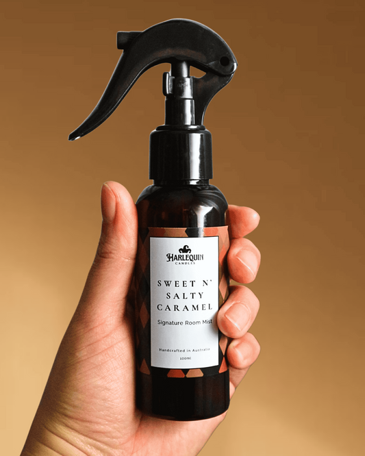 A Harlequin Candles Salted Caramel Signature Room Mist in an amber spray bottle with a black trigger cap, placed against a two-toned background of light yellow and brown. The label clearly shows 'Harlequin Candles', 'Sweet N' Salty Caramel', 'Signature Room Mist', and 'Handcrafted in Australia', highlighting this enchanting Australian-made home spray.