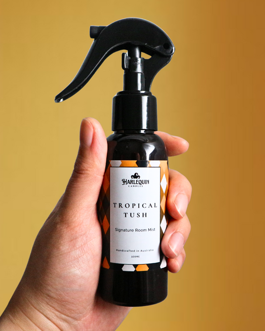A Harlequin Candles (Scented like popular Brazilian bum bum creams) Tropical Tush Signature Room Mist in an amber spray bottle with a black trigger cap, placed against a two-toned background of light blue and white. The label clearly shows 'Harlequin Candles', 'Tropical Tush', 'Signature Room Mist', and 'Handcrafted in Australia', highlighting this elegant Australian-made home spray.