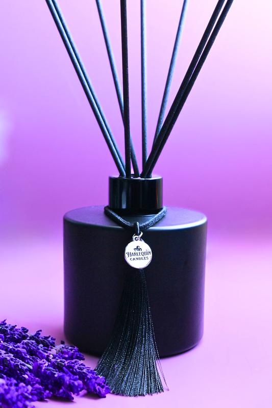 Harlequin Candles Vanilla Lavender Love Affair reed diffuser in a matte black bottle with black reeds and a decorative black tassel with a silver logo charm. A sprig of purple lavender lies beside the diffuser on a vibrant purple background, evoking a soothing and elegant home fragrance.