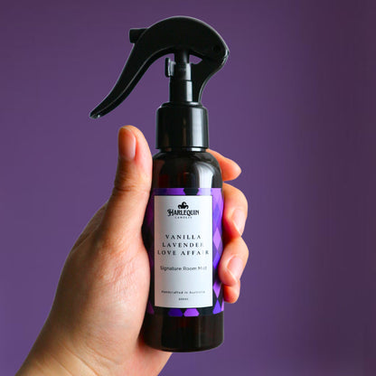 A Harlequin Candles Vanilla Lavender Love Affair Signature Room Mist in an amber spray bottle with a black trigger cap, placed against a two-toned background of light pink and white. The label clearly shows 'Harlequin Candles', 'Vanilla Lavender Love Affair', 'Signature Room Mist', and 'Handcrafted in Australia', highlighting this calming Australian-made home spray.