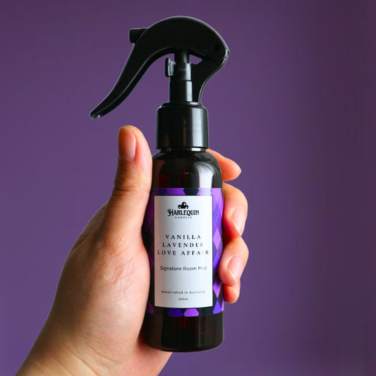 A Harlequin Candles Vanilla Lavender Love Affair Signature Room Mist in an amber spray bottle with a black trigger cap, placed against a two-toned background of light pink and white. The label clearly shows 'Harlequin Candles', 'Vanilla Lavender Love Affair', 'Signature Room Mist', and 'Handcrafted in Australia', highlighting this calming Australian-made home spray.