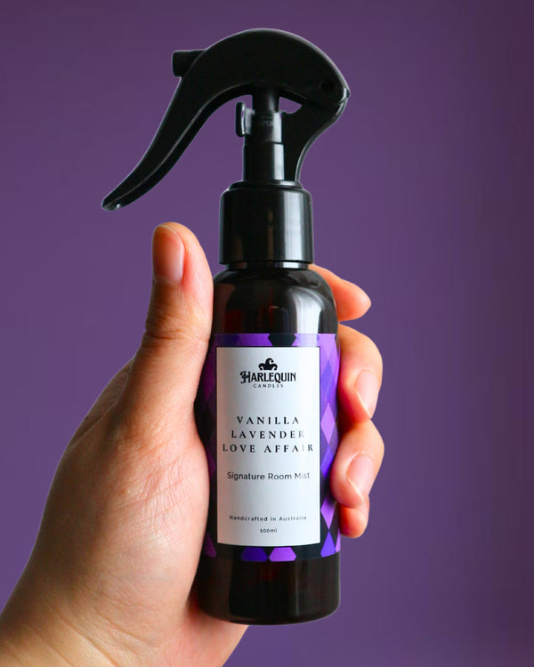 A Harlequin Candles Vanilla Lavender Love Affair Signature Room Mist in an amber spray bottle with a black trigger cap, placed against a two-toned background of light pink and white. The label clearly shows 'Harlequin Candles', 'Vanilla Lavender Love Affair', 'Signature Room Mist', and 'Handcrafted in Australia', highlighting this calming Australian-made home spray.