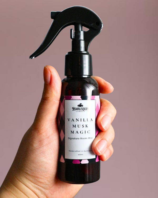 A Harlequin Candles Vanilla Musk Magic Signature Room Mist in an amber spray bottle with a black trigger cap, placed against a two-toned background of light pink and white. The label clearly shows 'Harlequin Candles', 'Vanilla Musk Magic', 'Signature Room Mist', and 'Handcrafted in Australia', highlighting this enchanting Australian-made home spray.