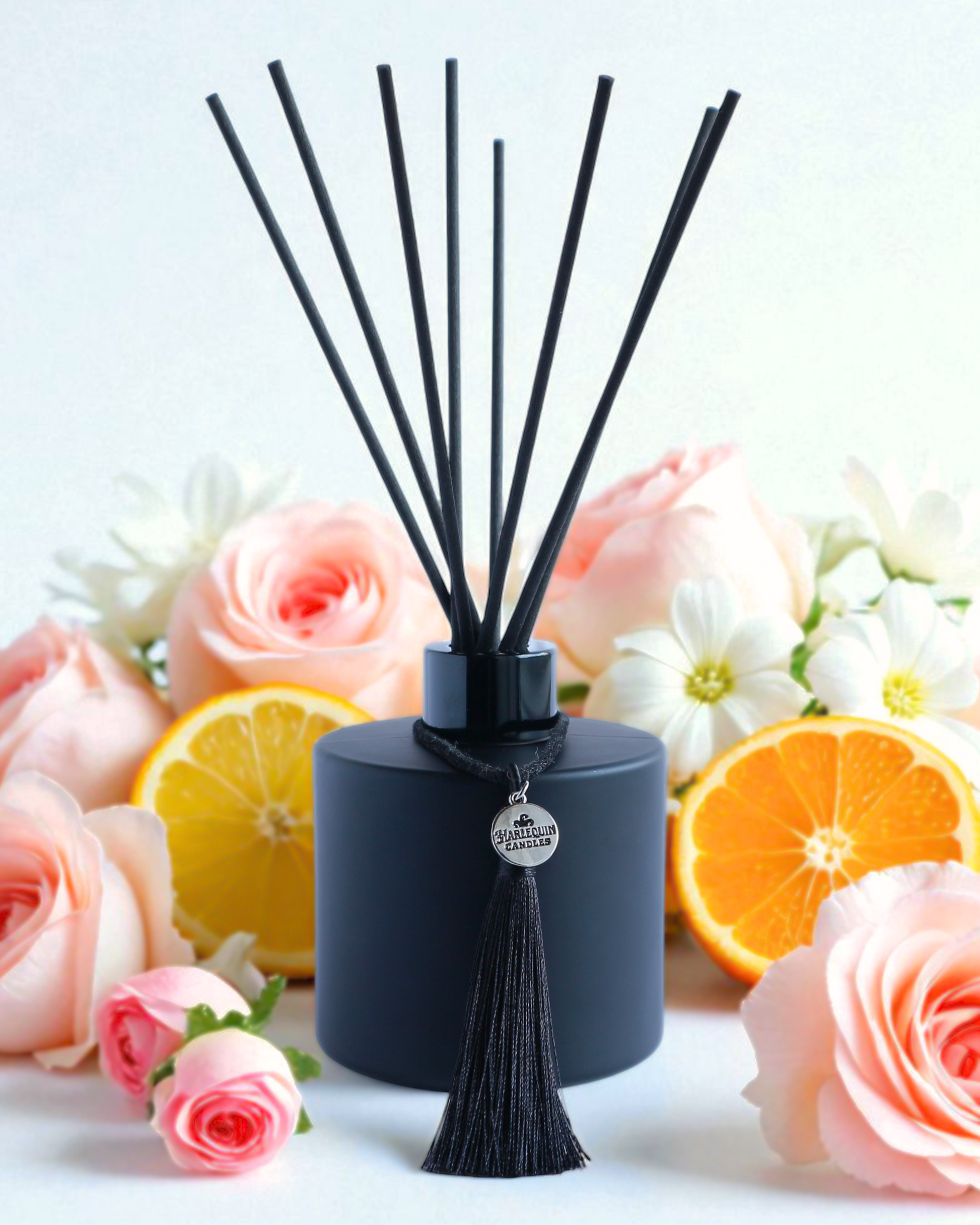 Harlequin Candles Vanilla Musk reed diffuser in a matte black bottle with a decorative black tassel and silver logo charm. A bundle of black reeds and several vanilla beans are arranged nearby, suggesting a warm and sweet marshmallow-infused home fragrance.
