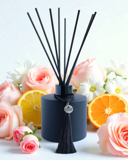Harlequin Candles Vanilla Musk reed diffuser in a matte black bottle with a decorative black tassel and silver logo charm. A bundle of black reeds and several vanilla beans are arranged nearby, suggesting a warm and sweet marshmallow-infused home fragrance.