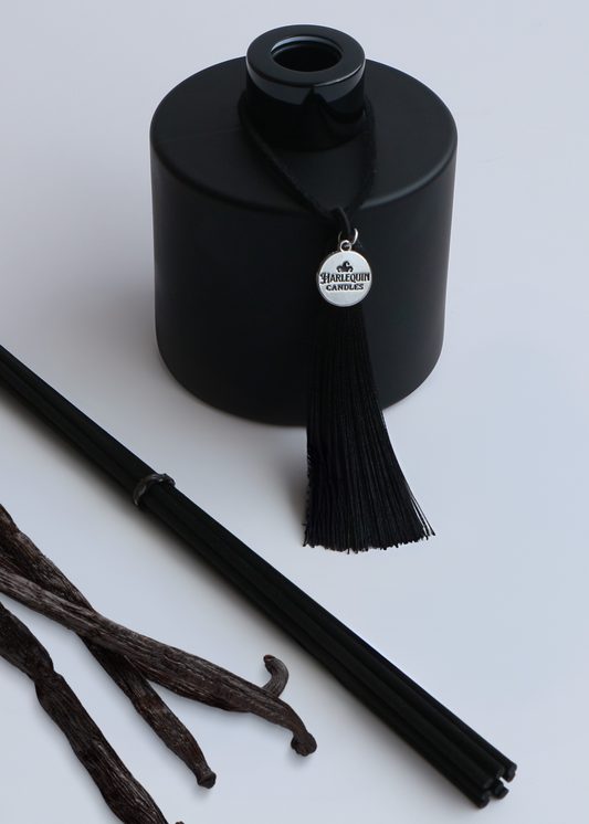 Harlequin Candles Vanilla Musk reed diffuser in a matte black bottle with a decorative black tassel and silver logo charm. A bundle of black reeds and several vanilla beans are arranged nearby, suggesting a warm and sweet marshmallow-infused home fragrance.