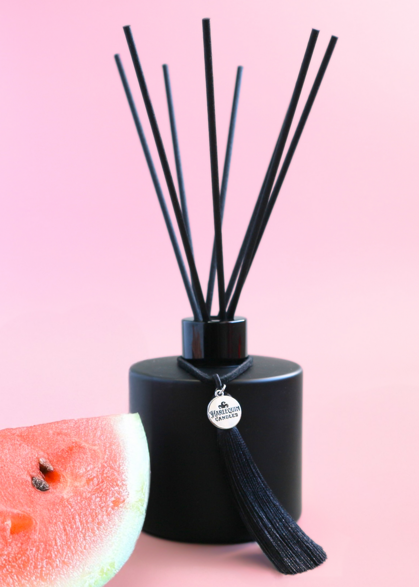 Harlequin Candles Watermelon Lemonade reed diffuser in a matte black bottle with black reeds and a decorative black tassel with a silver logo charm. A large pink flower is blurred in the foreground, set against a vibrant pink background, evoking a sweet and refreshing home aroma.
