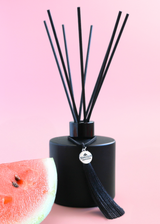 Harlequin Candles Watermelon Lemonade reed diffuser in a matte black bottle with black reeds and a decorative black tassel with a silver logo charm. A large pink flower is blurred in the foreground, set against a vibrant pink background, evoking a sweet and refreshing home aroma.