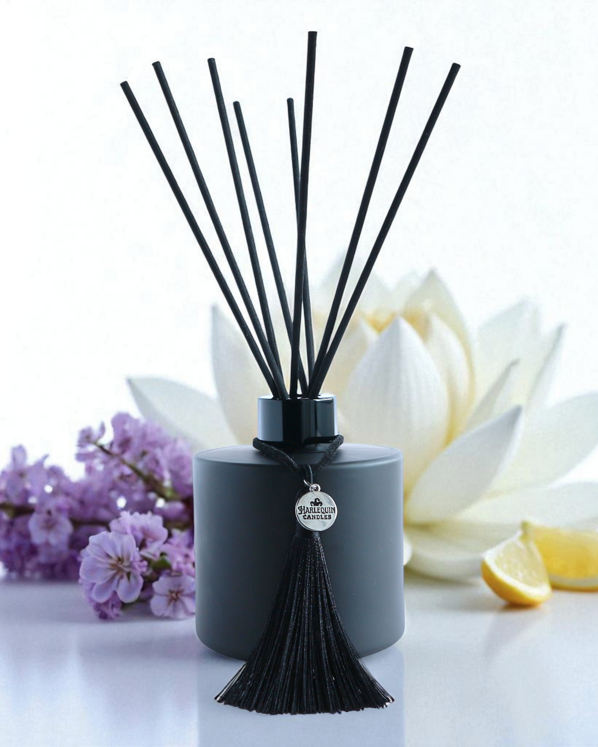 Harlequin Candles White Lotus reed diffuser in a matte black bottle with black reeds and a decorative black tassel with a silver logo charm. It stands on a round white display stand, flanked by large white lotus-like flowers in the background, conveying a serene and elegant home fragrance.