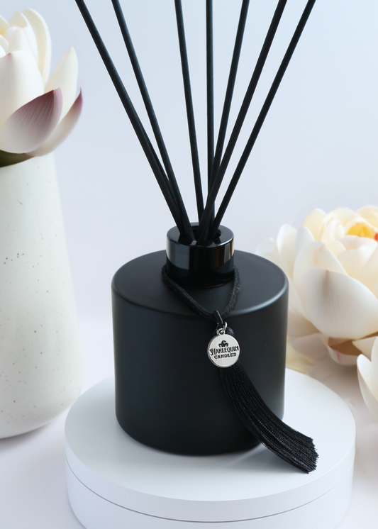 Harlequin Candles White Lotus reed diffuser in a matte black bottle with black reeds and a decorative black tassel with a silver logo charm. It stands on a round white display stand, flanked by large white lotus-like flowers in the background, conveying a serene and elegant home fragrance.