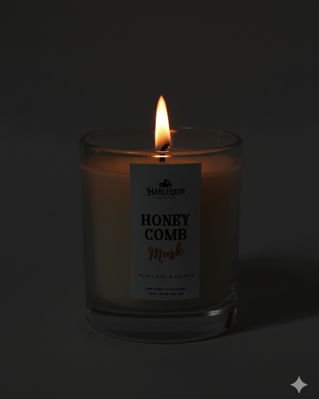 Lit Harlequin Candles Honeycomb Musk candle in a dark glass jar against a dark background, emphasizing the warm, cozy glow of the flame. Describes a proudly Australian-made, hand-poured soy wax candle.