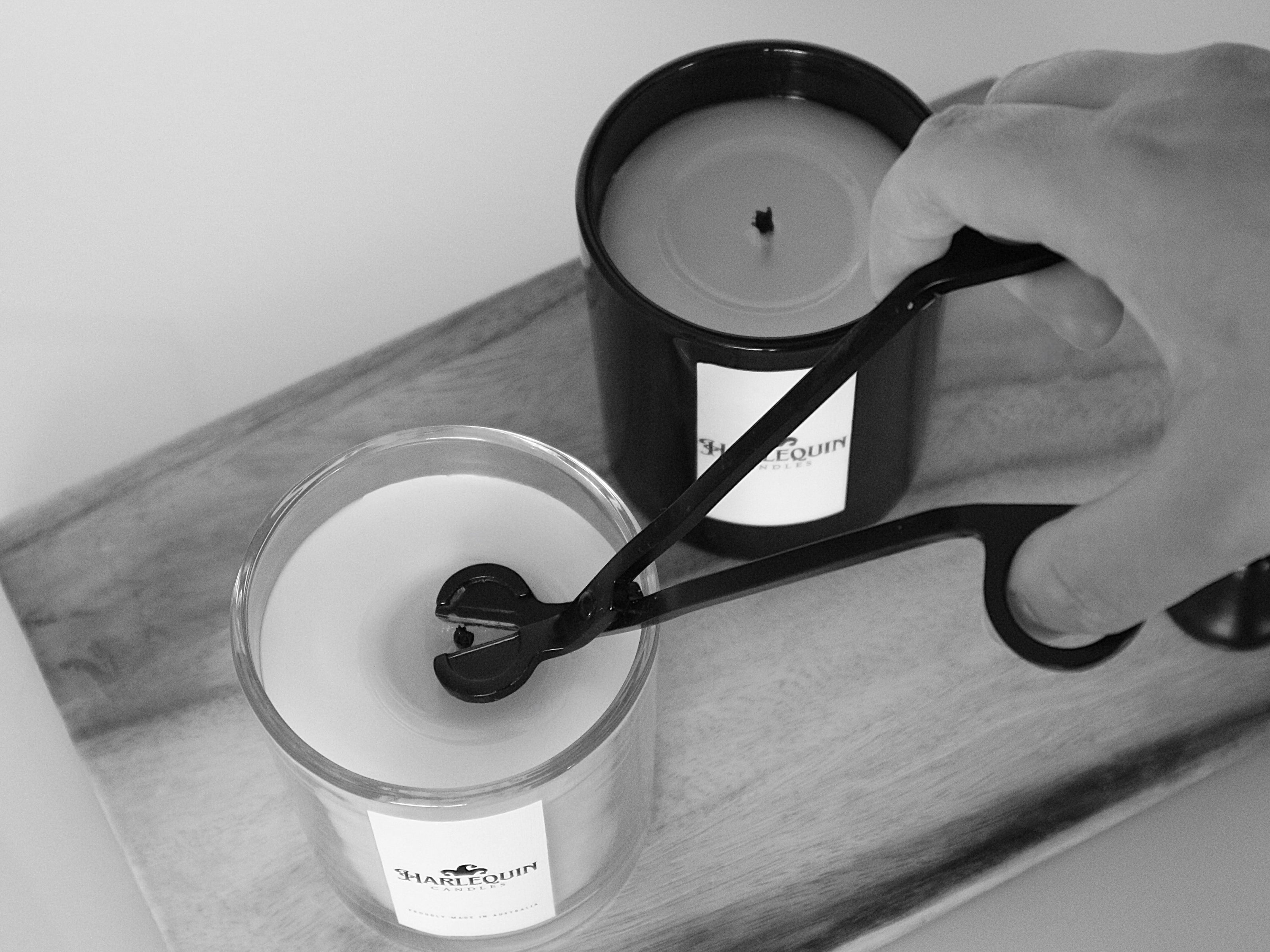 A hand uses a black candle wick trimmer to precisely trim the wick of a Harlequin Candles soy candle in a clear glass jar. Another Harlequin Candles candle in a black jar is visible behind it, all resting on a wooden board. Illustrates essential candle care for preventing soot and ensuring a longer, cleaner burn.