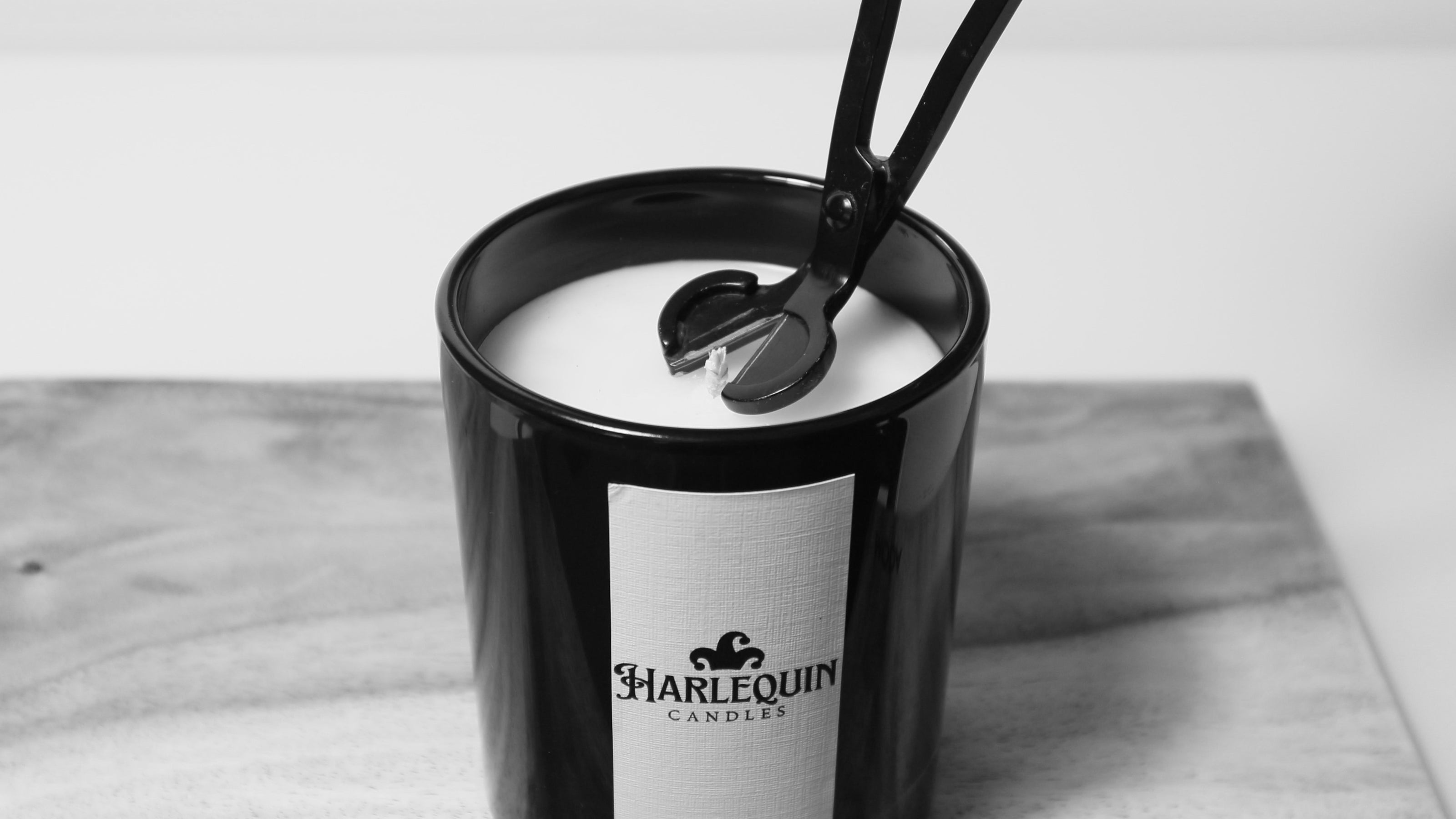 Close-up of a black candle wick trimmer positioned precisely over the wick of an unlit Harlequin Candles soy candle in a matte black glass jar. The candle rests on a wooden board, illustrating the essential step of wick trimming for a clean, even, and long-lasting burn.