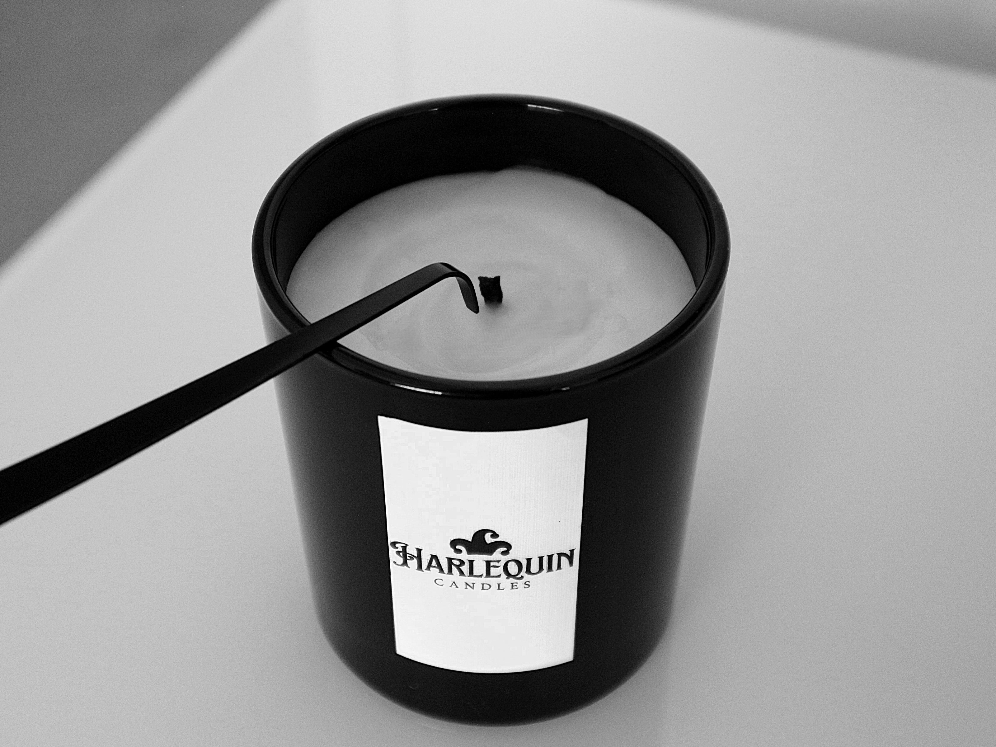 Monochrome close-up of a black wick dipper gently pushing the burning wick of a Harlequin Candles soy candle into its melted wax pool in a matte black glass jar. Illustrates the best way to extinguish a candle without smoke, ensuring cleaner candle care.