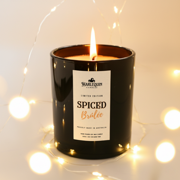 Black jar handmade soy candle labeled 'Spiced Brulee' by Harlequin Candles with a blurred light background. Limited edition Christmas candle.