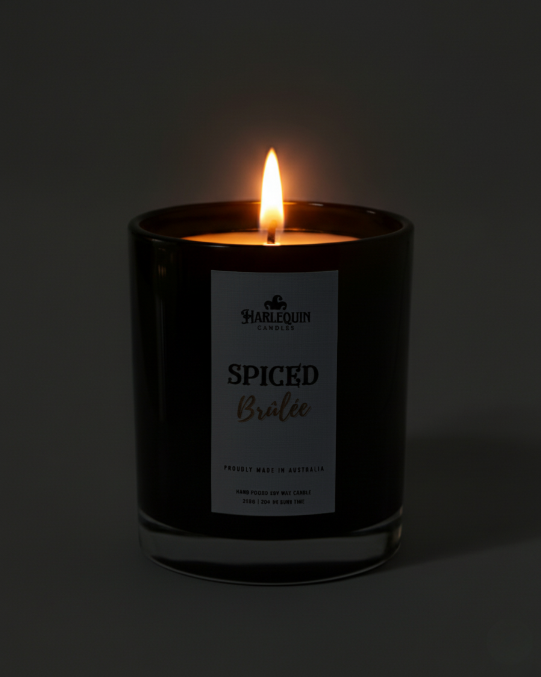 Lit Harlequin Candles Creme Brulee candle in a dark glass jar against a dark background, emphasizing the warm, cozy glow of the flame. Describes a proudly Australian-made, hand-poured soy wax candle.