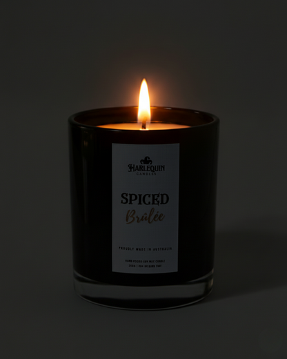Lit Harlequin Candles Creme Brulee candle in a dark glass jar against a dark background, emphasizing the warm, cozy glow of the flame. Describes a proudly Australian-made, hand-poured soy wax candle.