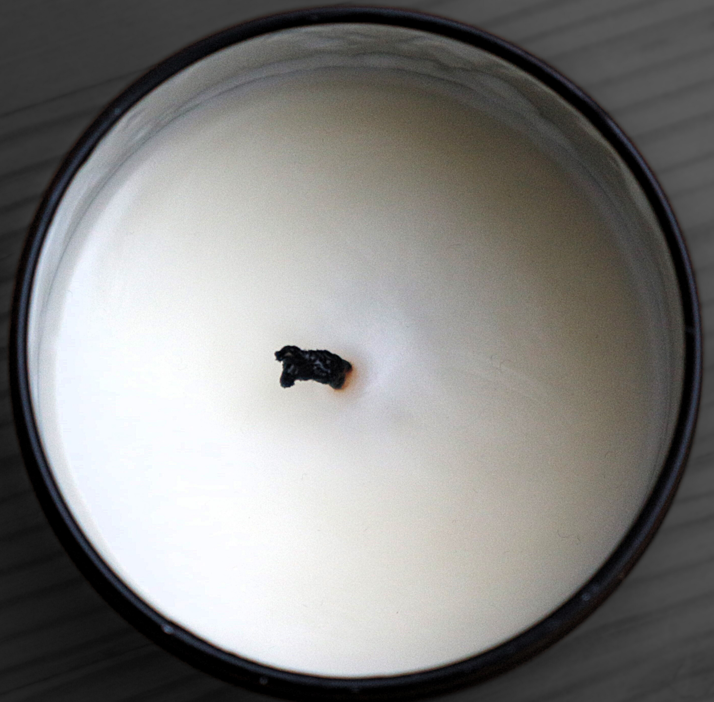 Overhead close-up of an unlit Harlequin Candles soy candle in a dark glass jar, showing a black, mushroomed wick with carbon buildup. Illustrates the result of an untrimmed wick and the importance of proper candle care for a clean burn.