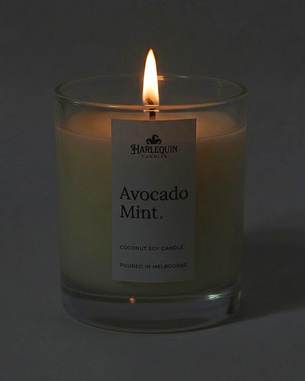 A lit Harlequin Candles Avocado Mint coconut soy candle in a clear glass jar. The warm, flickering flame creates a soft glow against the minimalist white label, which features the brand logo and the text "Poured in Melbourne" against a dark, atmospheric background.