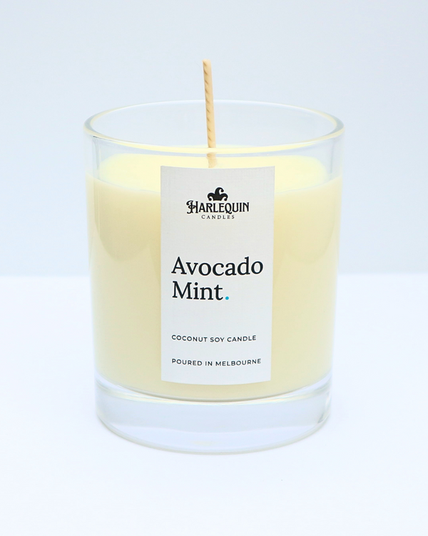 A clear glass jar filled with a creamy pale yellow coconut soy candle labeled Avocado Mint by Harlequin Candles. The label features a minimalist design and specifies the candle was poured in Melbourne.