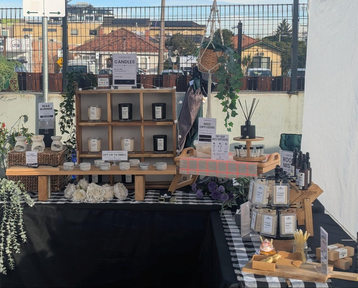 A market stall display for 'Handmade Home Fragrance' products under a white canopy banner that reads 'Proudly Made in Melbourne'. The stall features wooden shelves with small bottles, jars, and decorative items, alongside a black draped table and a checkered table with candles and reeds.