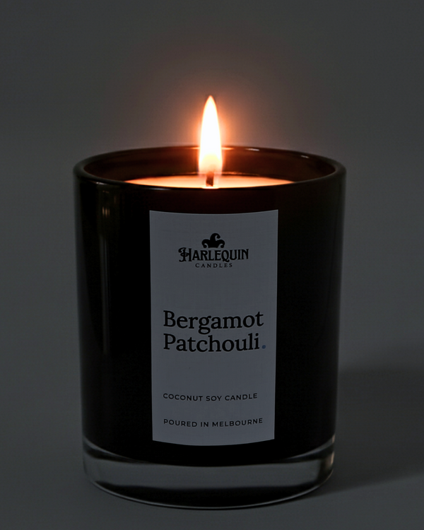 A lit Harlequin Candles Bergamot Patchouli coconut soy candle in a glossy black glass jar. The warm flame illuminates the minimalist white label, which features bold black typography and the text "Poured in Melbourne" against a dark, moody background.