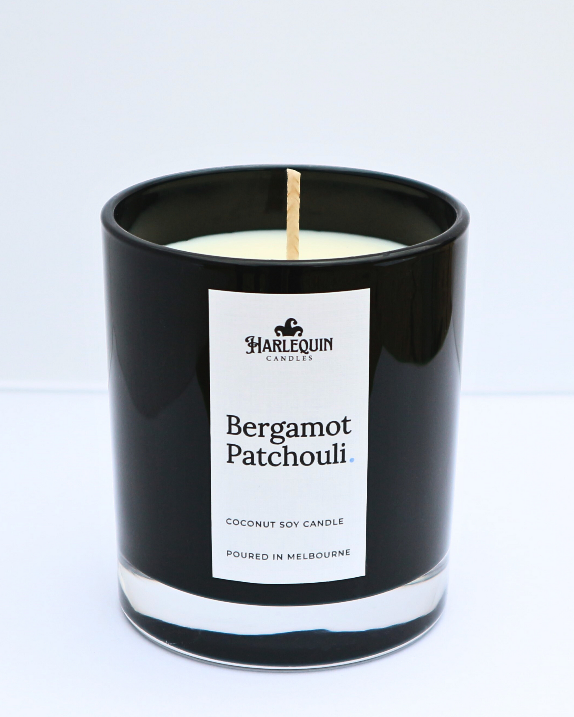 A sleek, glossy black glass jar containing a Bergamot Patchouli coconut soy candle by Harlequin Candles. The minimalist white label features the brand's logo and the text "Poured in Melbourne" at the bottom, set against a bright, clean background.