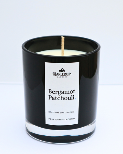 A sleek, glossy black glass jar containing a Bergamot Patchouli coconut soy candle by Harlequin Candles. The minimalist white label features the brand's logo and the text "Poured in Melbourne" at the bottom, set against a bright, clean background.