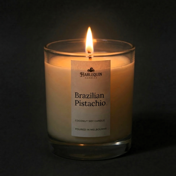 A lit Brazilian Pistachio coconut soy candle by Harlequin Candles in a clear glass jar. The warm flame illuminates the minimalist white label, which features the brand logo and the text "Poured in Melbourne" against a dark, atmospheric background.