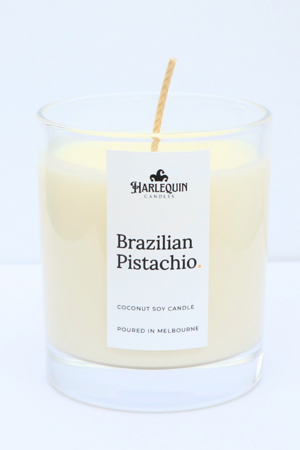 A bright, clear glass jar containing a Brazilian Pistachio coconut soy candle by Harlequin Candles. The minimalist white label features the Harlequin logo and the text "Poured in Melbourne" at the base.