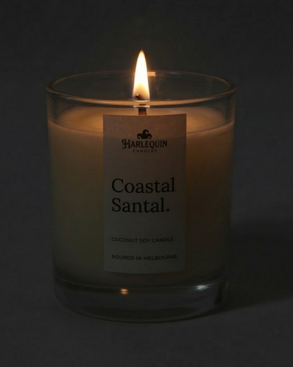 A lit Harlequin Candles luxury Coastal Santal coconut soy candle in a clear glass jar. The warm glow of the flame highlights the minimalist label and the premium wax, with the text "Poured in Melbourne" clearly visible at the bottom.