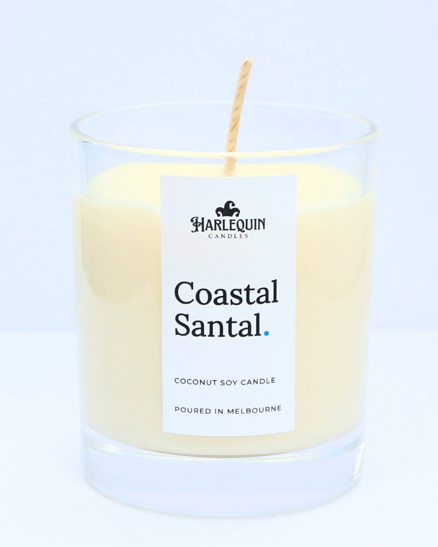 A close-up of a Coastal Santal coconut soy candle by Harlequin Candles in a clear glass jar. The minimalist white label features the brand's logo and the text "Poured in Melbourne" against a clean white background.