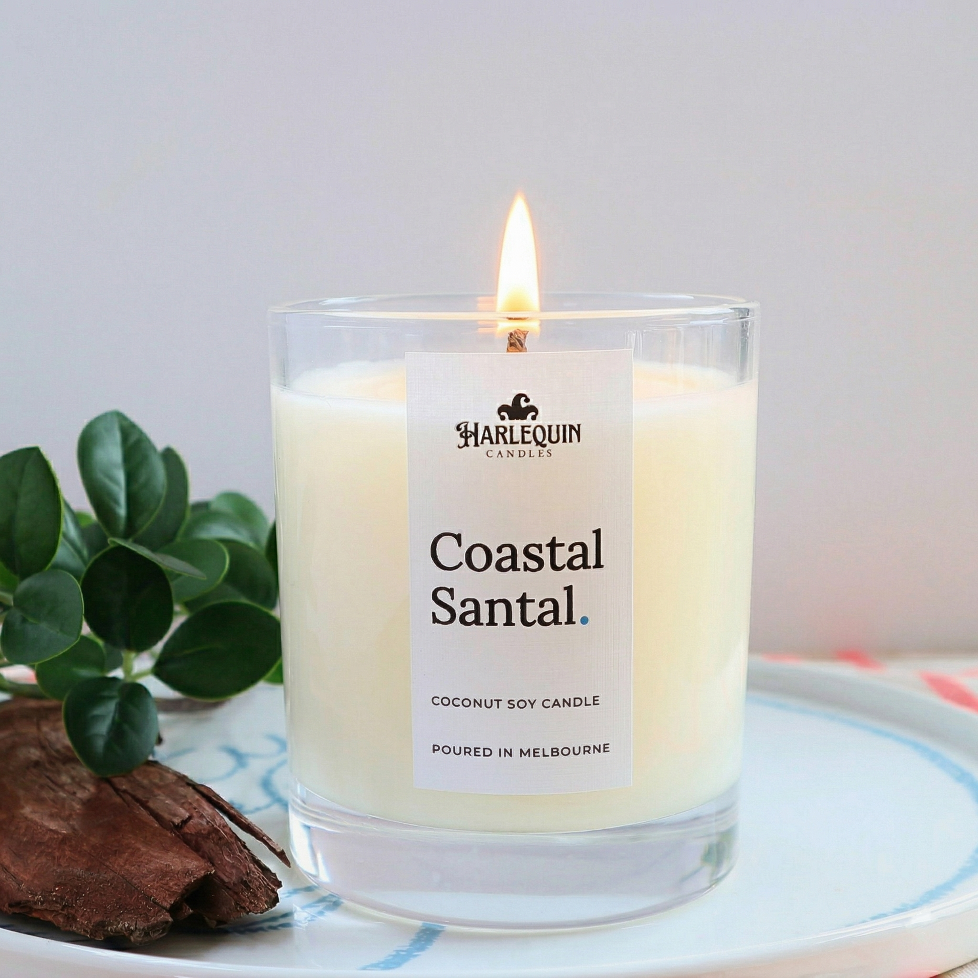 Harlequin Candles Coastal Santal coconut soy wax candle in a clear jar, featuring a minimalist label and styled with natural wood and green leaves.