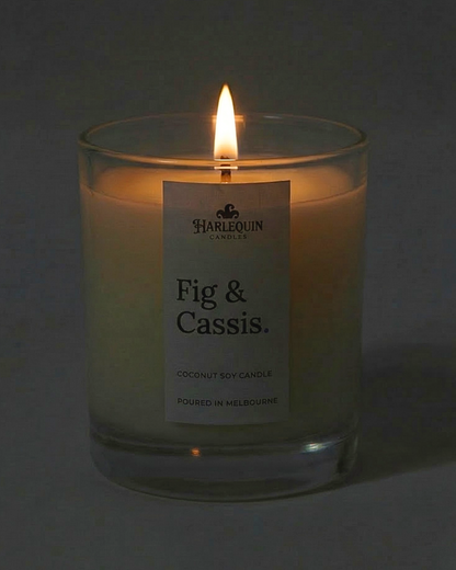 A lit Fig & Cassis coconut soy candle by Harlequin Candles in a clear glass jar. The warm flame illuminates the minimalist white label, which features the brand's logo and the text "Poured in Melbourne" against a soft, dark background.