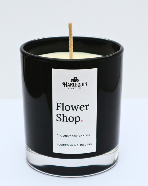 A sleek black glass jar containing a Flower Shop coconut soy candle by Harlequin Candles. The minimalist white label features bold black text and the brand's logo, with the text "Poured in Melbourne" at the bottom against a clean, bright background.