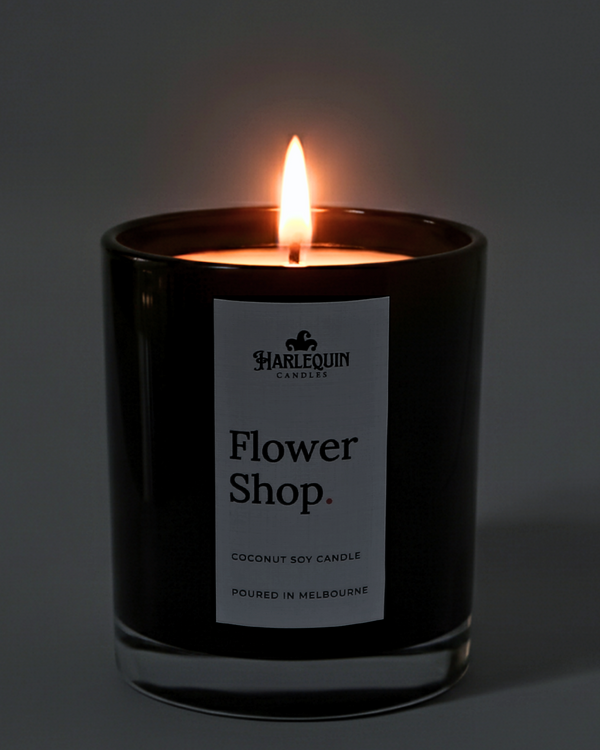 A lit Harlequin Candles luxury Flower Shop coconut soy candle in a glossy black glass jar. The warm flame illuminates the minimalist white label, which features bold typography and the text "Poured in Melbourne.