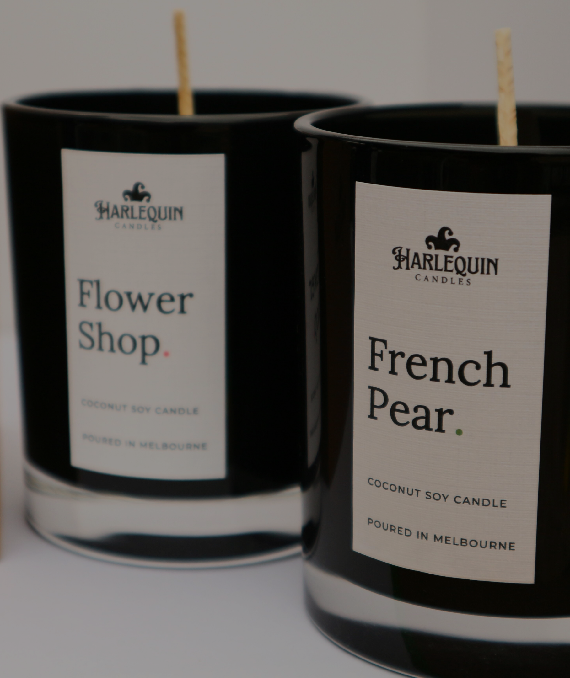 Two Harlequin Candles side-by-side in glossy black glass jars featuring the scents Flower Shop and French Pear. Both jars have minimalist white labels stating they are coconut soy candles and were poured in Melbourne.