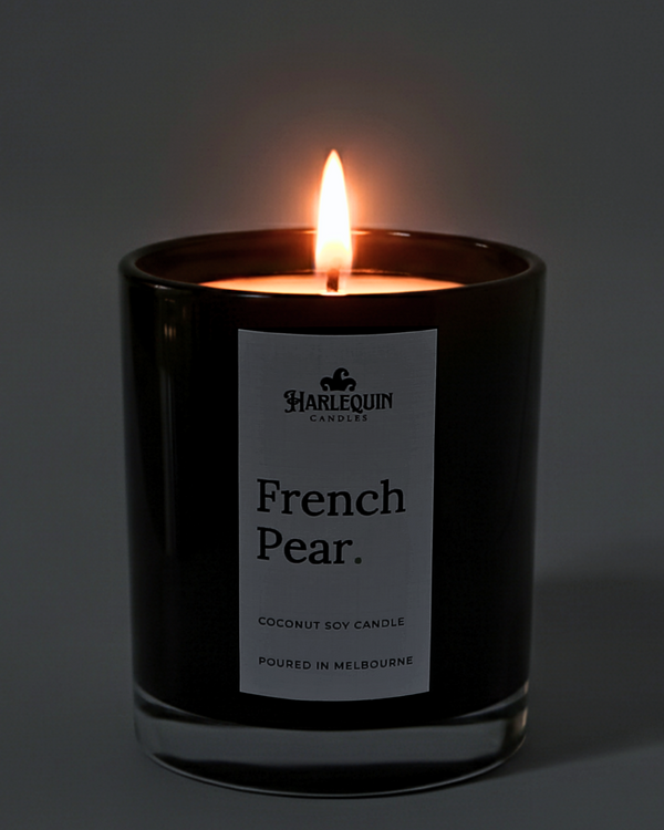 A lit Harlequin Candles premium French Pear coconut soy candle in a glossy black glass jar. The warm flame illuminates the minimalist white label, which features the brand logo and the text "Poured in Melbourne" against a soft, dark background.