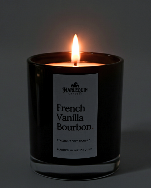 A lit Harlequin Candles luxury French Vanilla Bourbon coconut soy candle in a glossy black glass jar. The warm flame illuminates the minimalist white label, which features bold typography and the text "Poured in Melbourne.