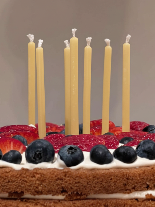 A close-up of eight tall, slender hand-dipped beeswax candles by Harlequin Candles. The natural yellow tapers are placed in a fresh berry cake with strawberries and blueberries. The image captures the unique, organic texture of the wax and the raw cotton wicks, emphasizing they are poured in Melbourne.
