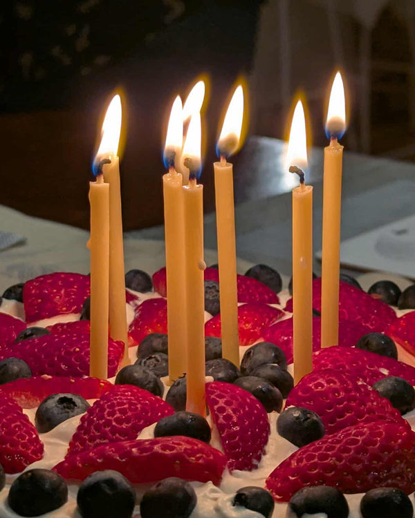 A set of six elegant, hand-dipped natural beeswax taper candles by Harlequin Candles. The slender yellow candles are lit, showing a soft, warm glow against a dark background, positioned as a premium topper for a fresh berry celebration cake.
