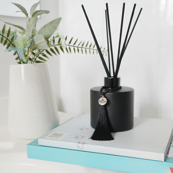 A luxury black reed diffuser, handmade in Australia by Harlequin Candles, featuring black fibre reeds and a decorative tassel, styled on decorative books.