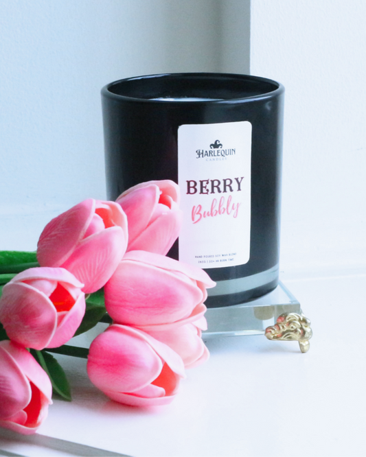 A Harlequin Candles "Berry Bubbly" hand-poured soy wax candle in a black glass jar, presented with a bouquet of pink tulips. Premium Australian made scented candle.