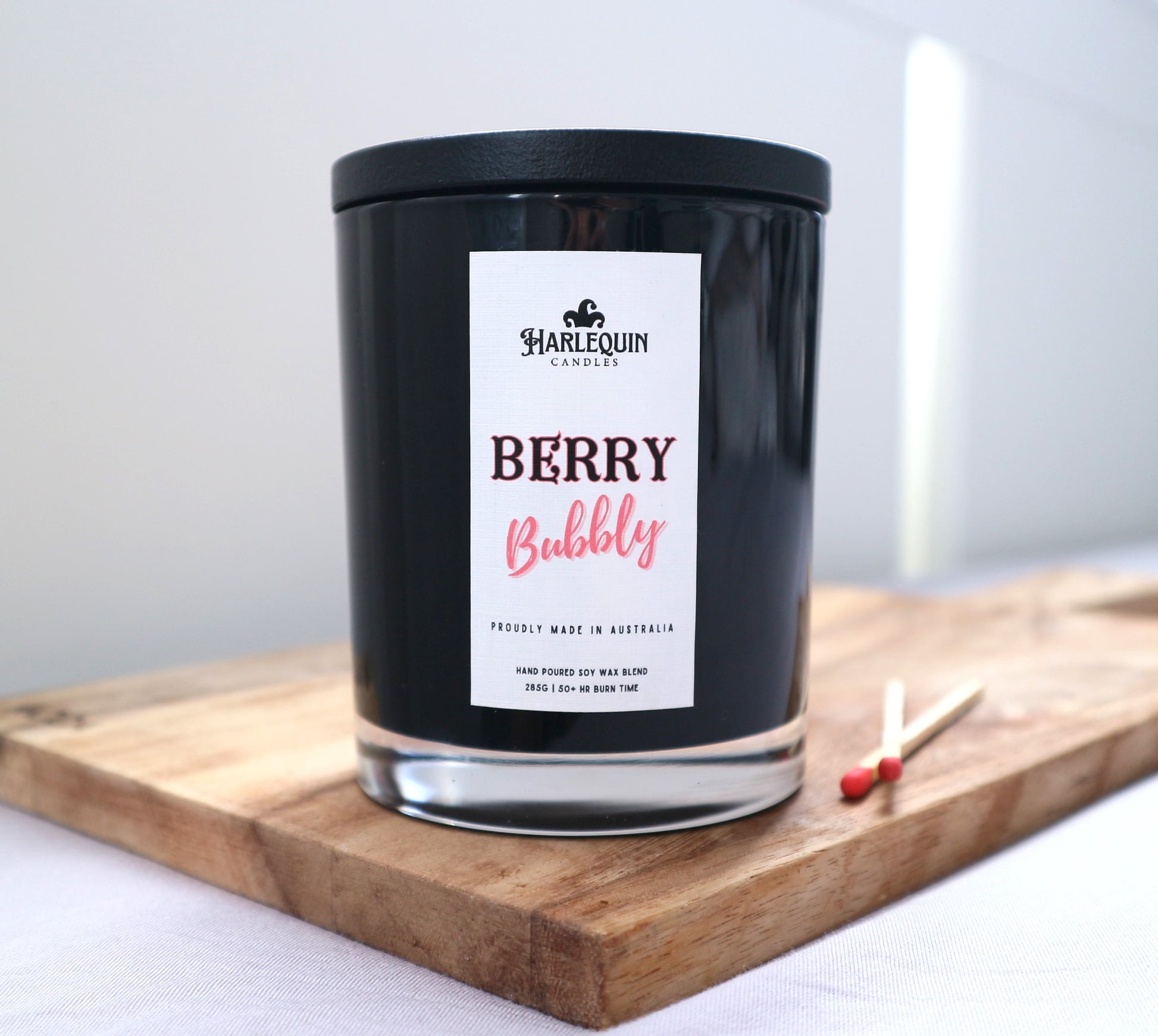 A Harlequin Candles "Berry Bubbly" hand-poured soy blend candle in a sleek black glass jar with lid, displayed on a wooden board with matches. Proudly Australian made.