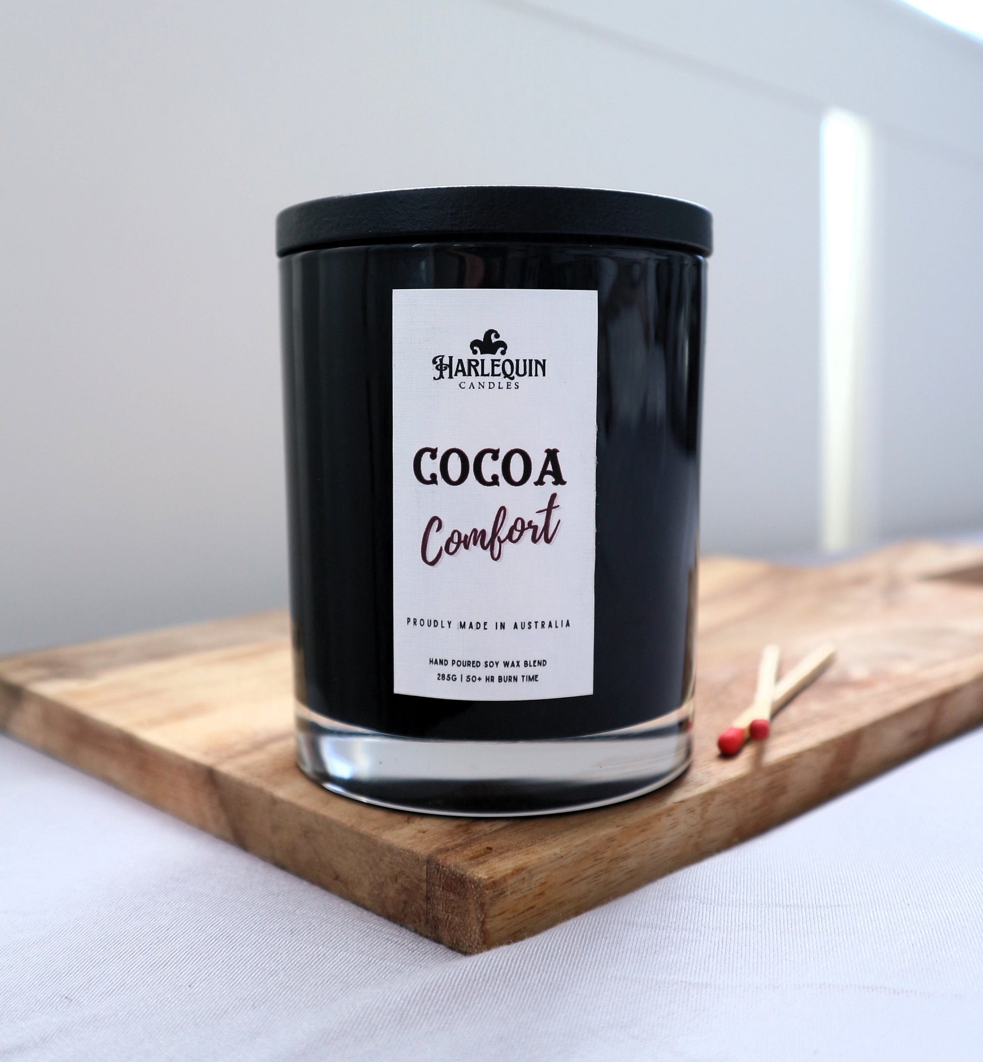 A Harlequin Candles "Cocoa Comfort" hand-poured soy blend candle in a sleek black glass jar with lid, displayed on a wooden board with matches. Proudly Australian made.