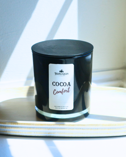 A Harlequin Candles "Cocoa Comfort" hand-poured soy wax candle in a black glass jar with lid, displayed on a light wooden tray in sunlight. Premium Australian made scented candle.