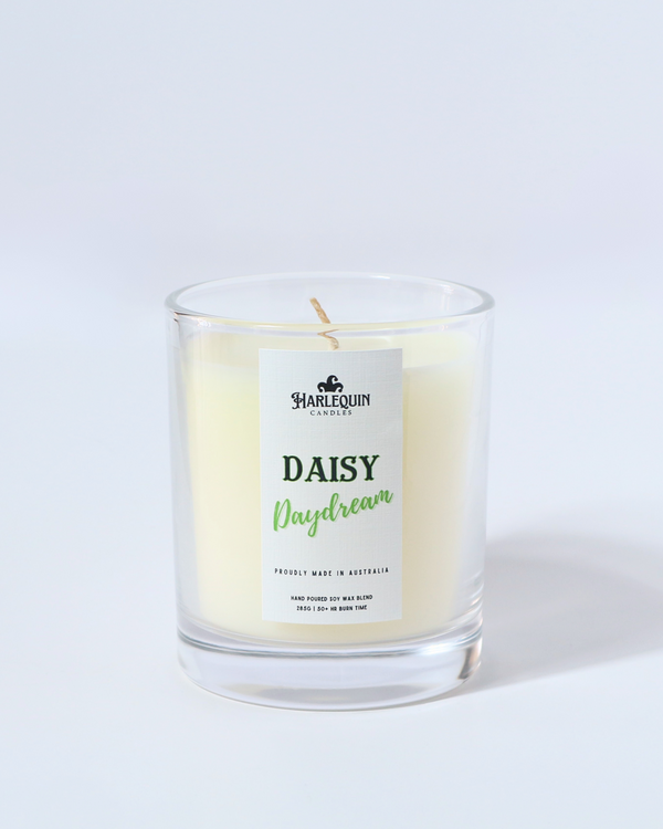 A Harlequin Candles "Daisy Daydream" hand-poured soy wax candle in a clear glass jar, displayed on a wooden pedestal with green plants in the background. Premium Australian made scented candle.